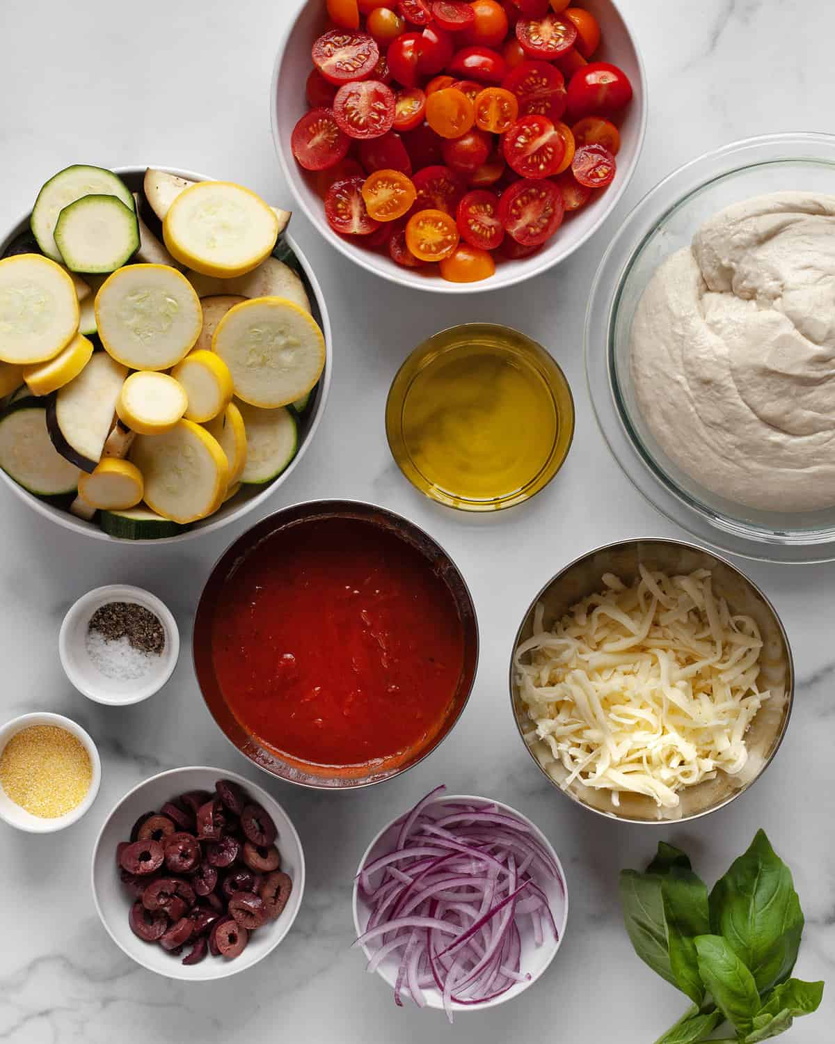 Ingredients including pizza dough in a bowl, vegetables, sauce, cheese, olives, onions, basil and olive oil.