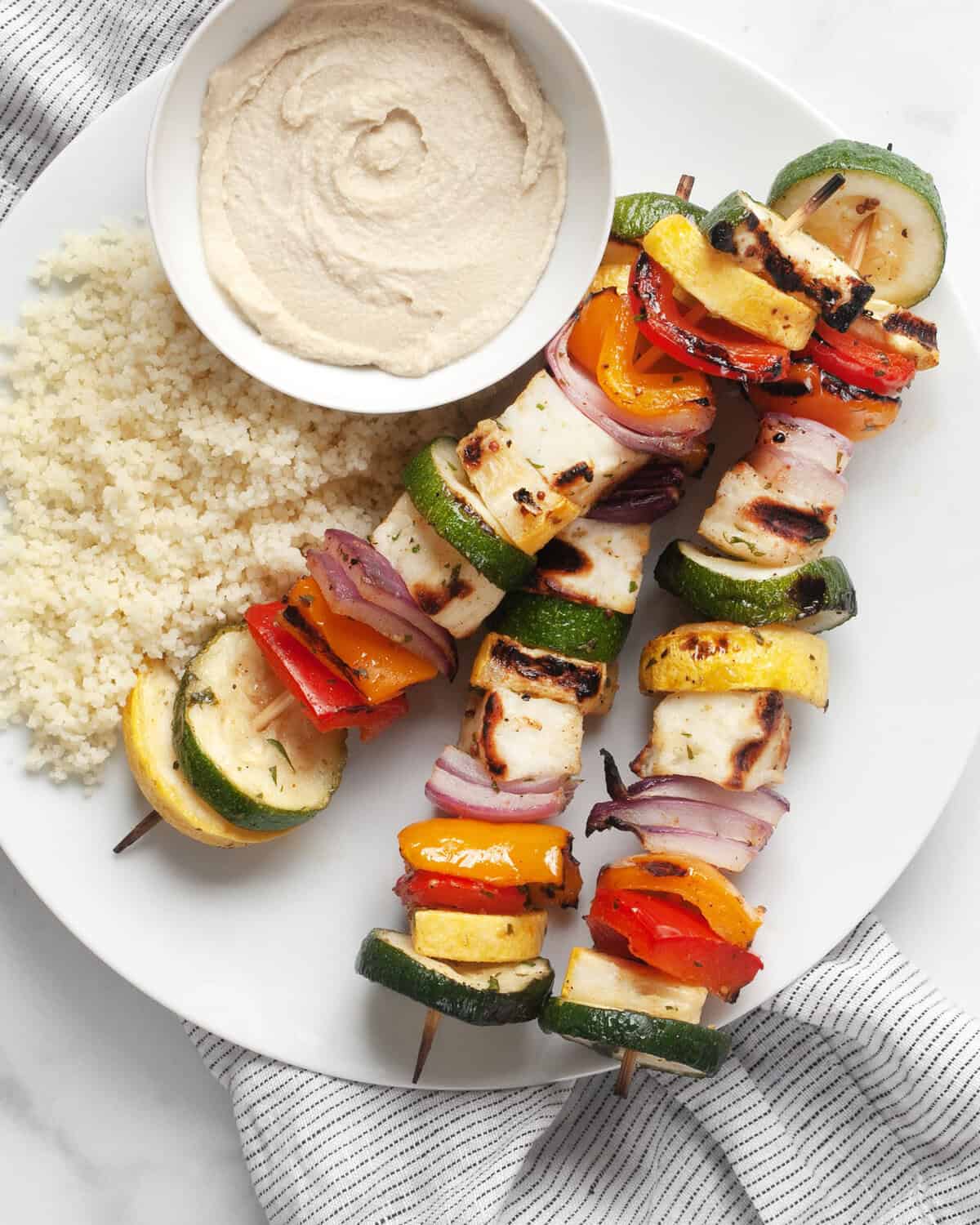 Three grilled veggie skewers on a plate with couscous and hummus.