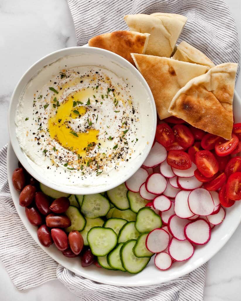 Labneh in a bowl with a platter of pita, tomatoes, radishes, cucumbers and olives