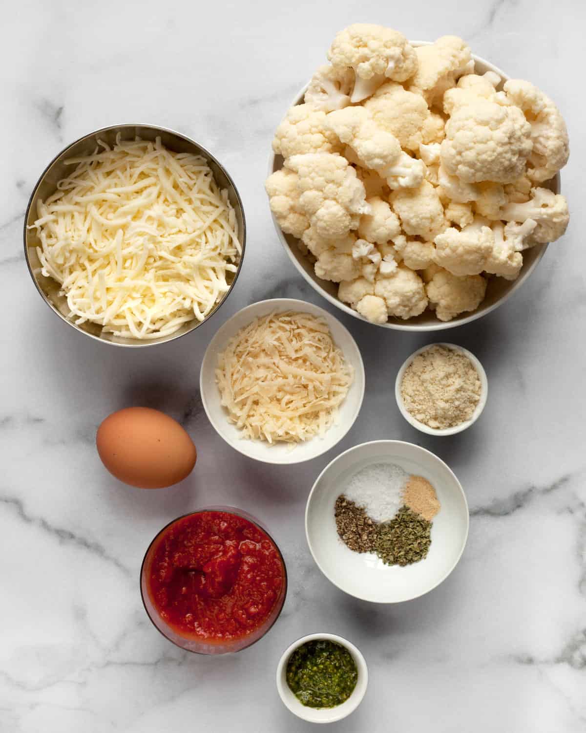 Ingredients including cauliflower, cheese, spices, almond flour, egg, pizza sauce and pesto.