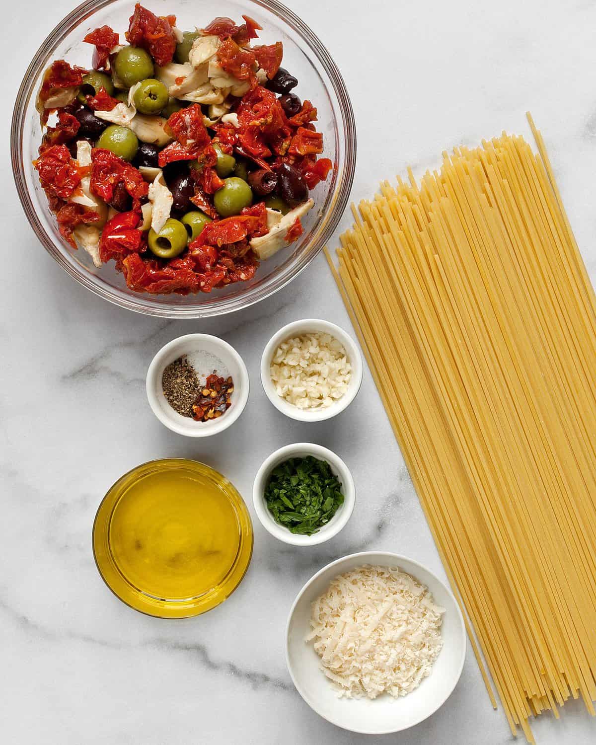 Ingredients including roasted tomatoes, olives, artichokes, spaghetti, parmesan, garlic and olive oil.