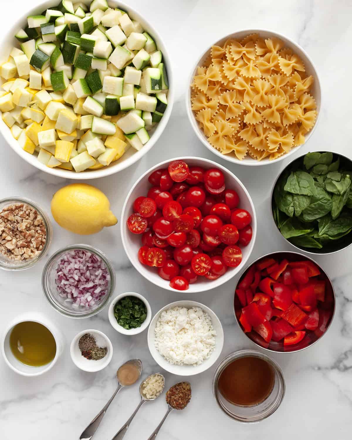 Ingredients including zucchini, yellow squash, bell peppers, tomatoes, bow tie pasta, baby spinach, red onions, walnuts, lemon, seasonings and olive oil.
