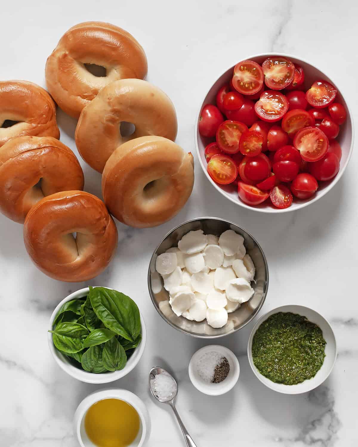 Ingredients including bagels, tomatoes, sliced mozzarella, pesto, basil, olive oil, kosher salt, black pepper and flaky sea salt.