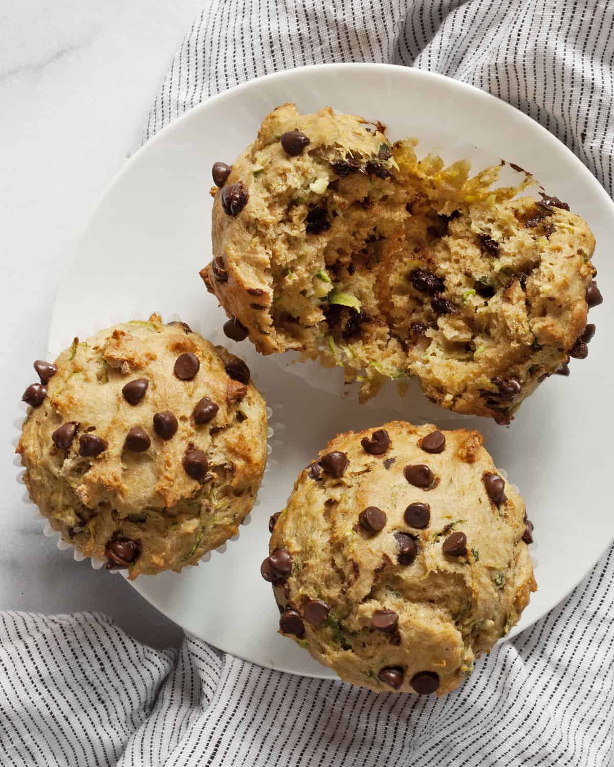 Three chocolate chip zucchini muffins on a plate with one torn in half.