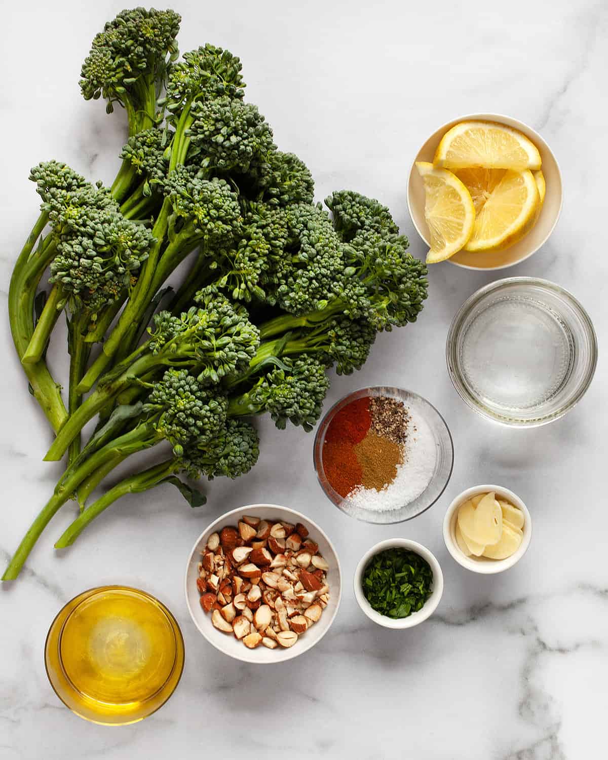 Ingredients including broccolini, olive oil, spices, almonds, garlic and lemon wedges.