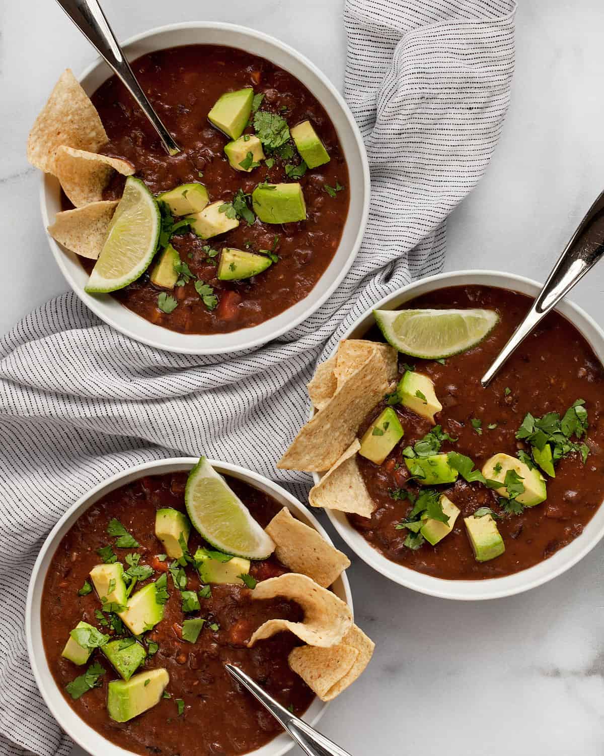 Three bowls of black bean soup topped with avocados and lime wedges.