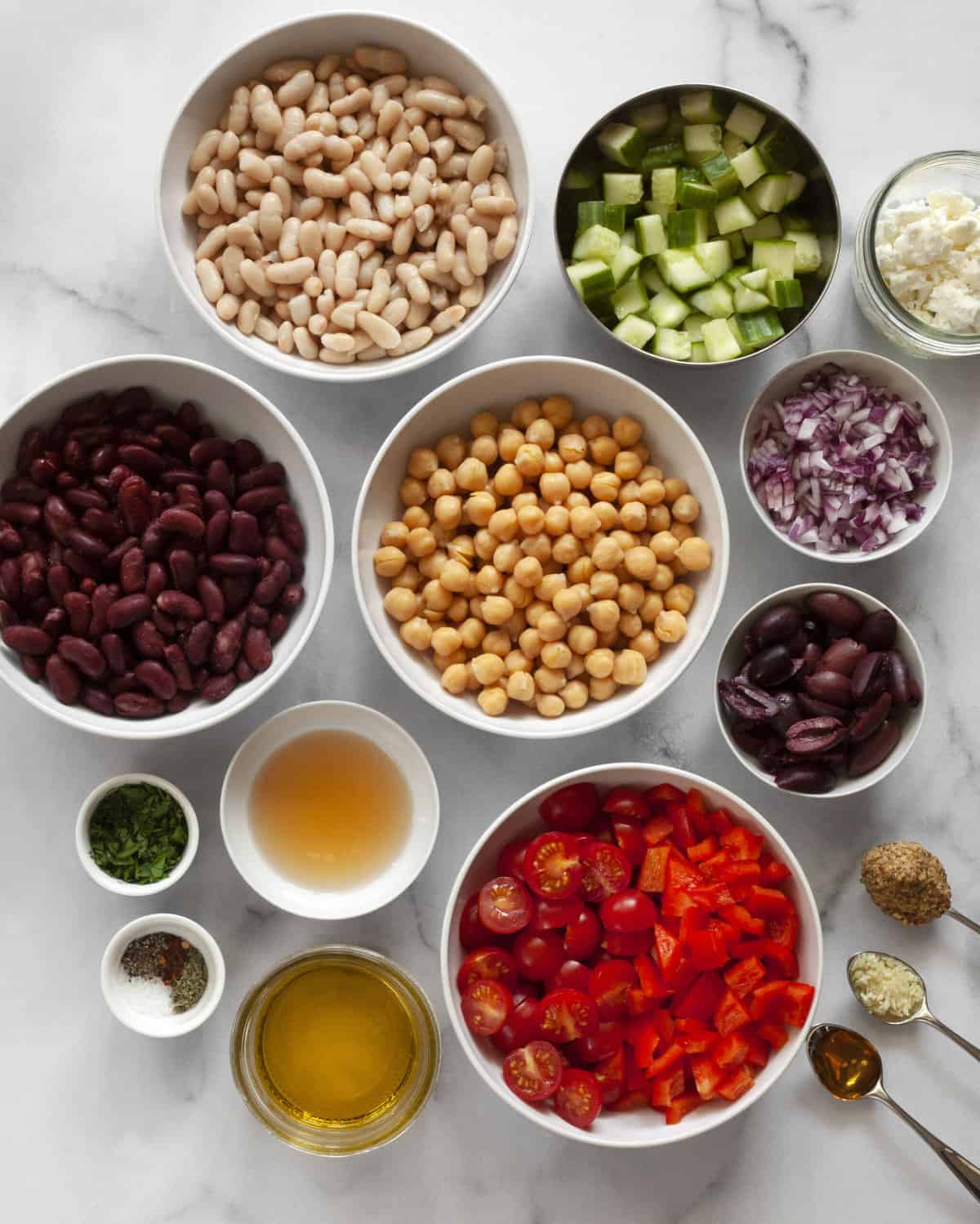 Ingredients including cannellini beans, kidney beans, chickpeas, red peppers, onions, cucumbers, vinegar, oil, spices, parsley and feta.