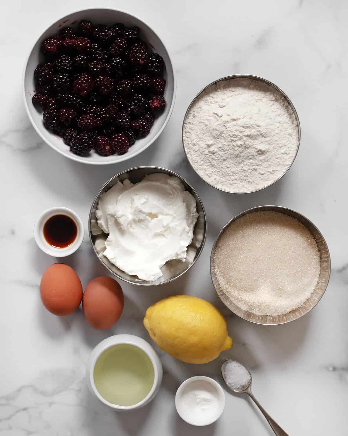 Ingredients including blackberries, flour, sugar, eggs, Greek yogurt, salt, baking powder, vanilla extract, lemon and oil.