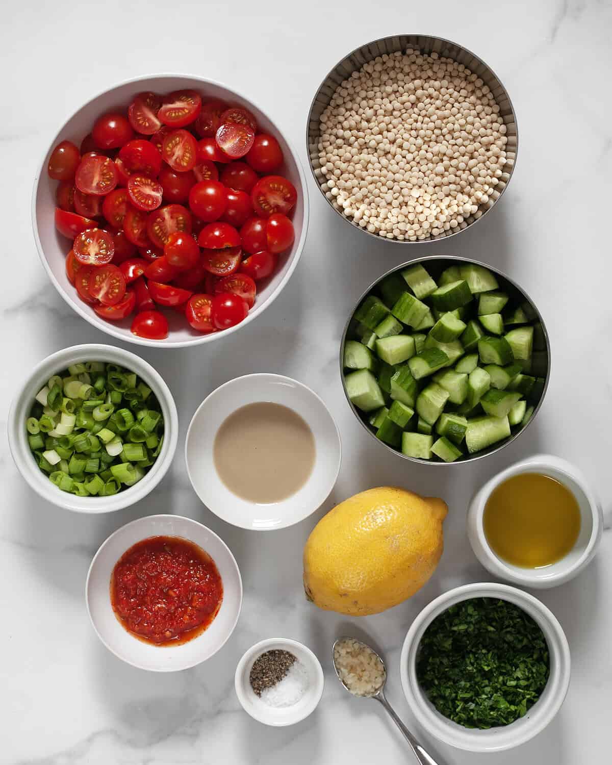 Ingredients including tomatoes, cucumbers, couscous, chopped parsley, scallions, lemon, tahini, harissa, garlic, salt, pepper and olive oil.