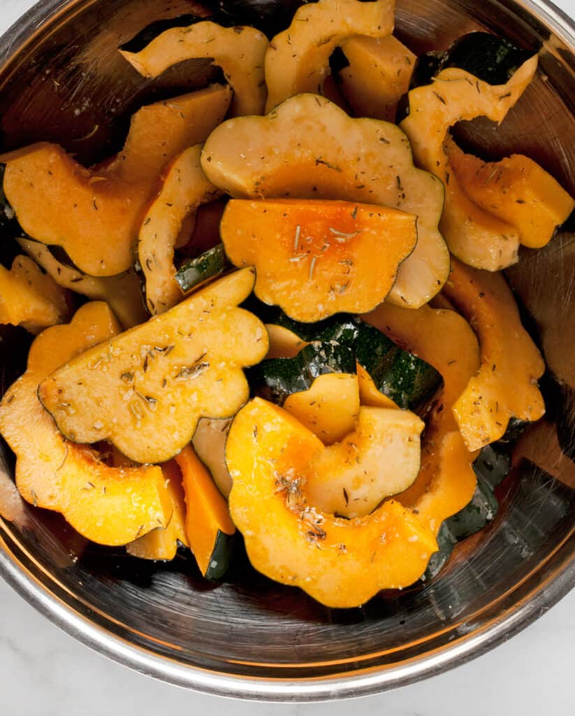 Sliced acorn squash in a bowl with olive oil and seasonings