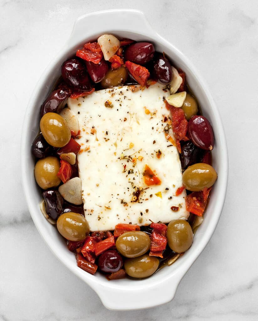 Block of feta in a dish with olives tomatoes, garlic and olive oil before it goes in the oven