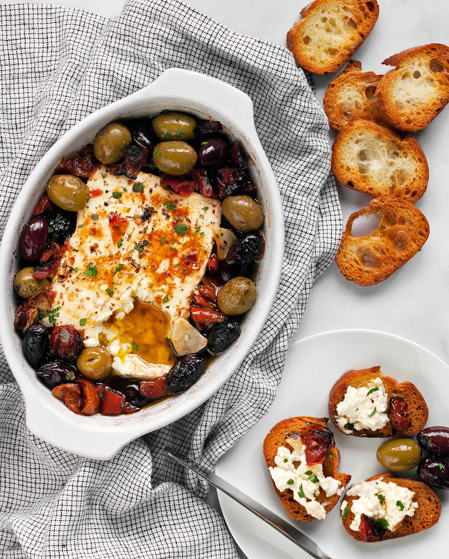 Baked feta with tomatoes and olive in a dish with toasted baguette slices.