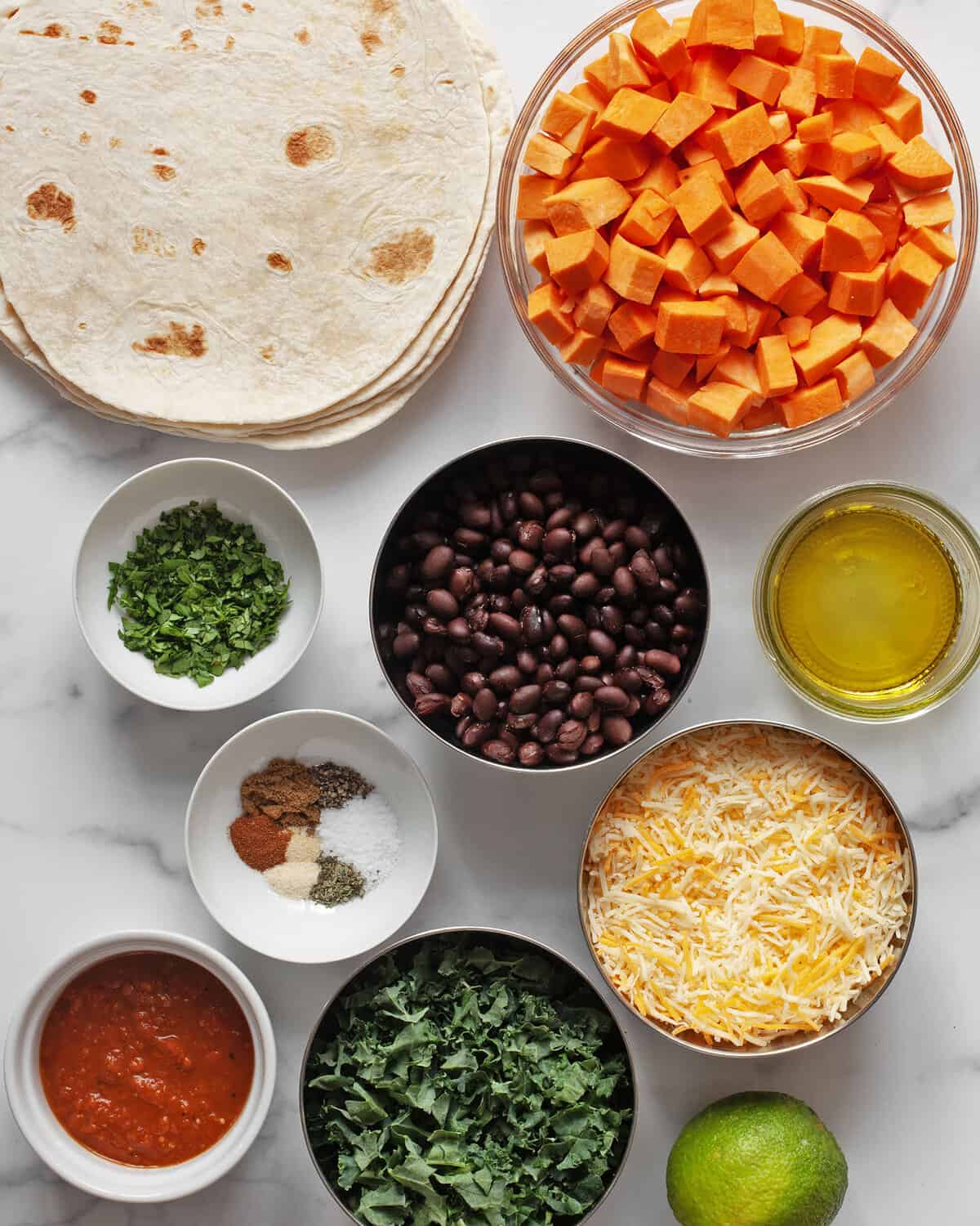 Ingredients including sweet potatoes, black beans, spices. tortillas, cheese, oil, cilantro, kale, salsa and lime.