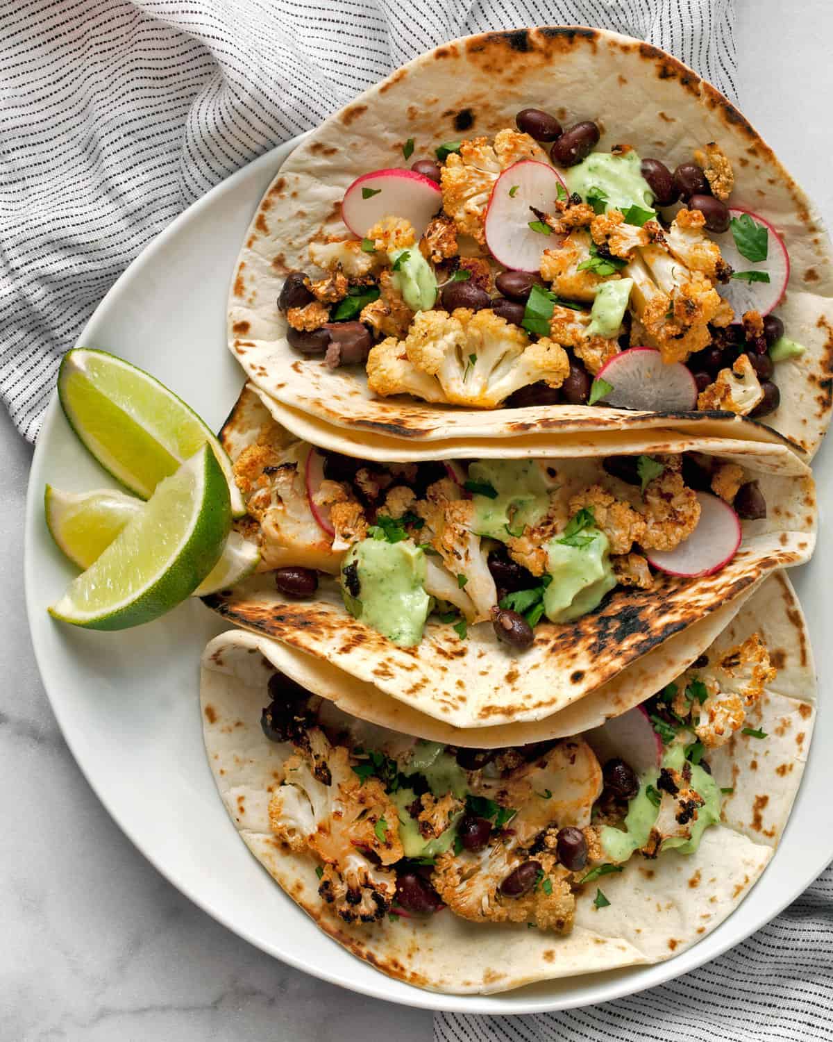 Three roasted vegetable tacos on a plate with slices of lime.