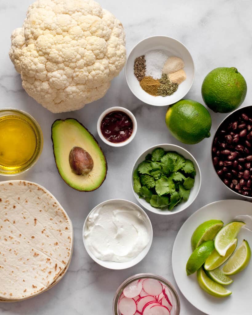 Ingredients including cauliflower, black beans, spices, limes, avocado, cilantro, yogurt and tortillas.