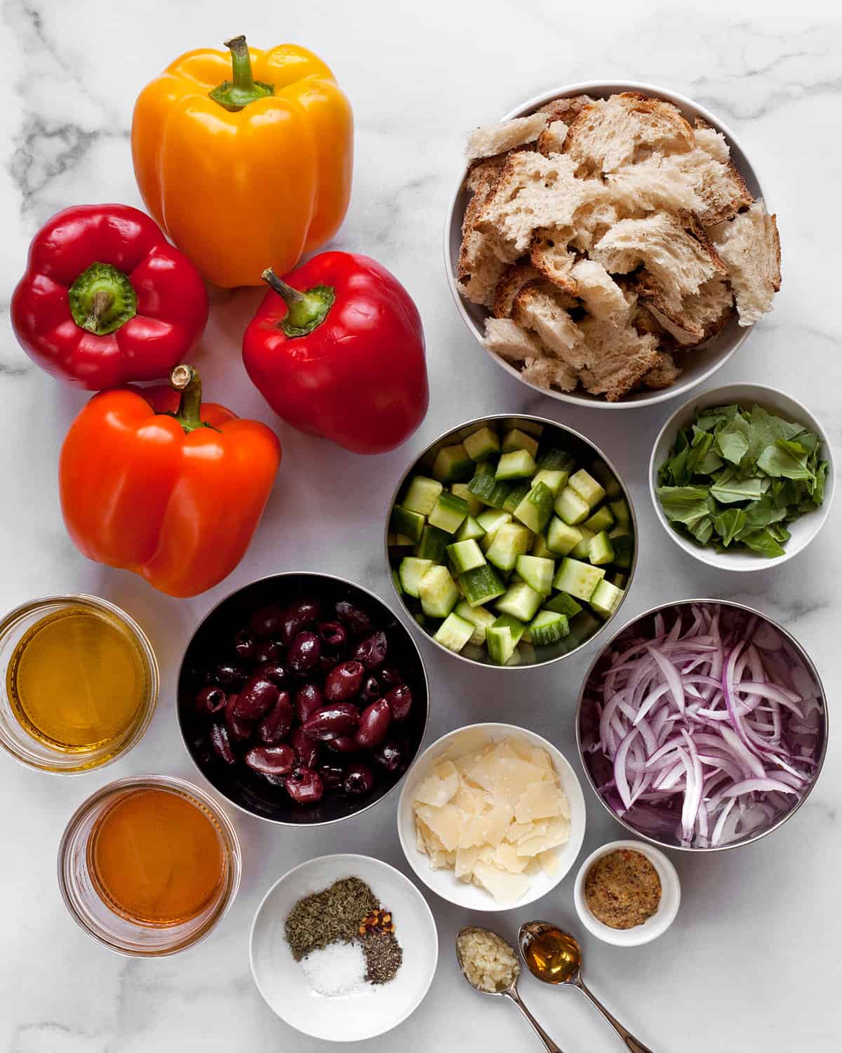 Ingredients including peppers, cucumbers, olives, red onions, Parmesan, bread, olive oil, seasonings, garlic, basil and vinegar.