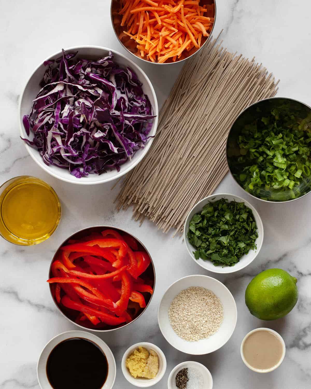 Ingredients including soba noodles, cabbage, carrots, bell peppers, scallions, cilantro, lime, sesame seeds, ginger, garlic and tahini.