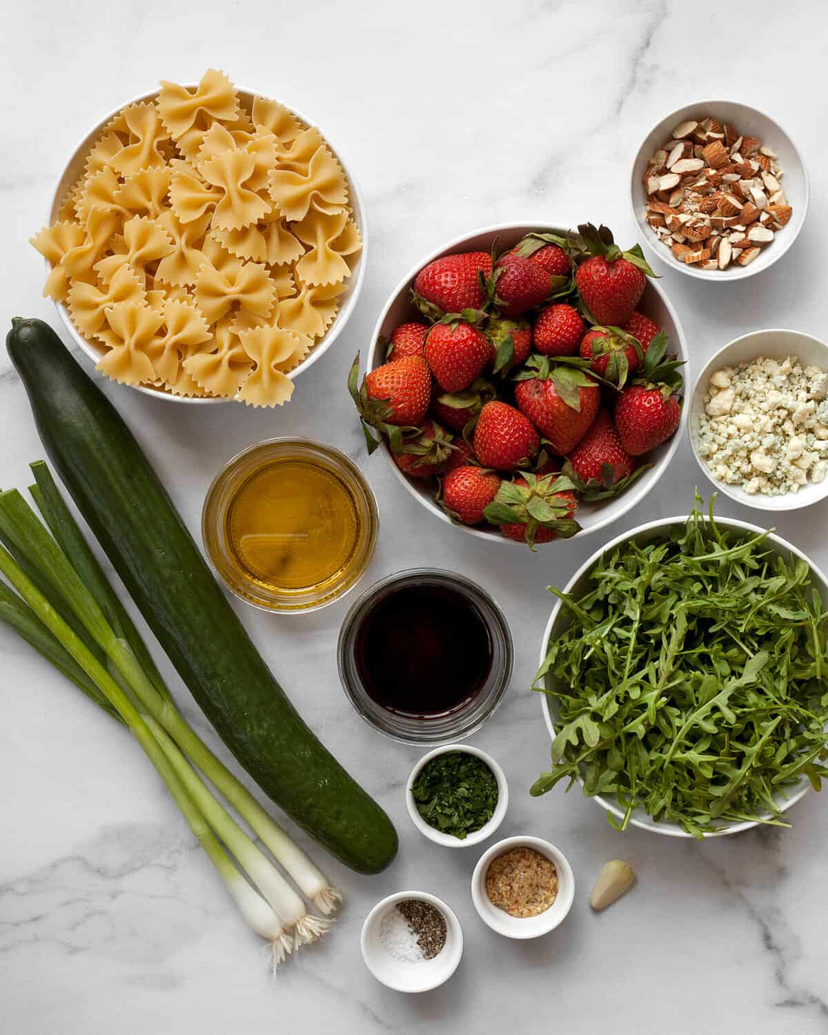 Ingredients including pasta, cucumber, strawberries, arugula, scallions, blue cheese, almonds, balsamic vinegar and olive oil.