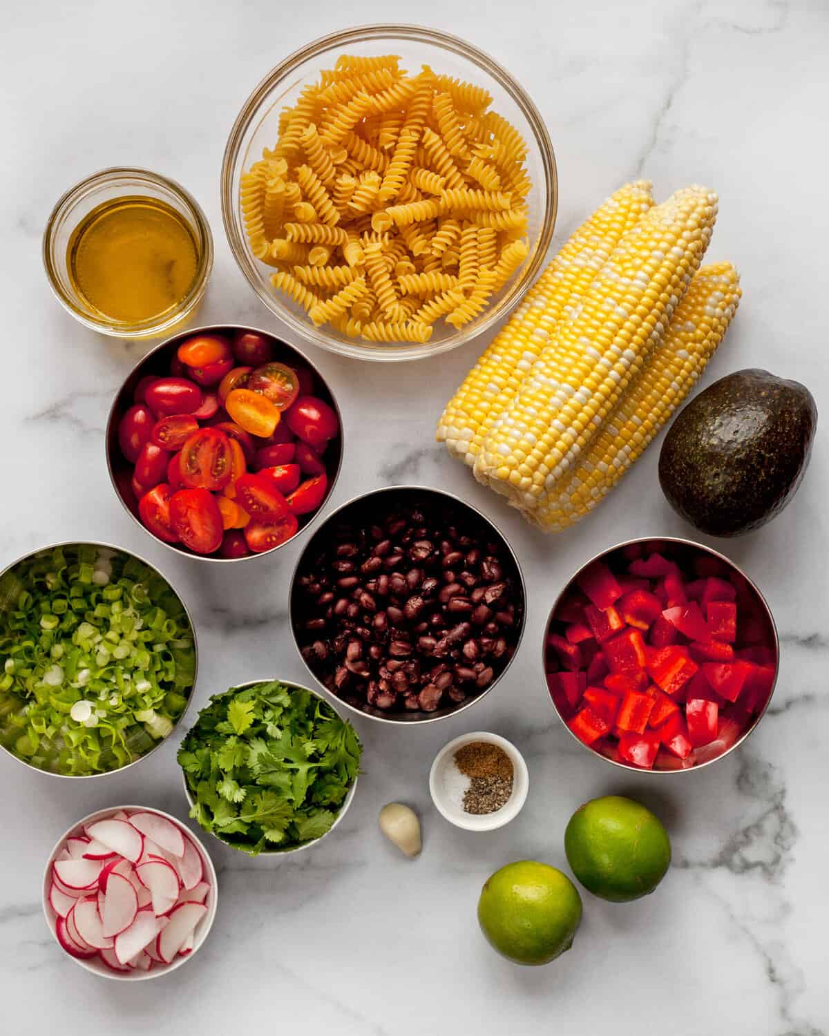 Ingredients including pasta, corn, tomatoes, peppers, black beans, avocado, lime, scallions and olive oil.