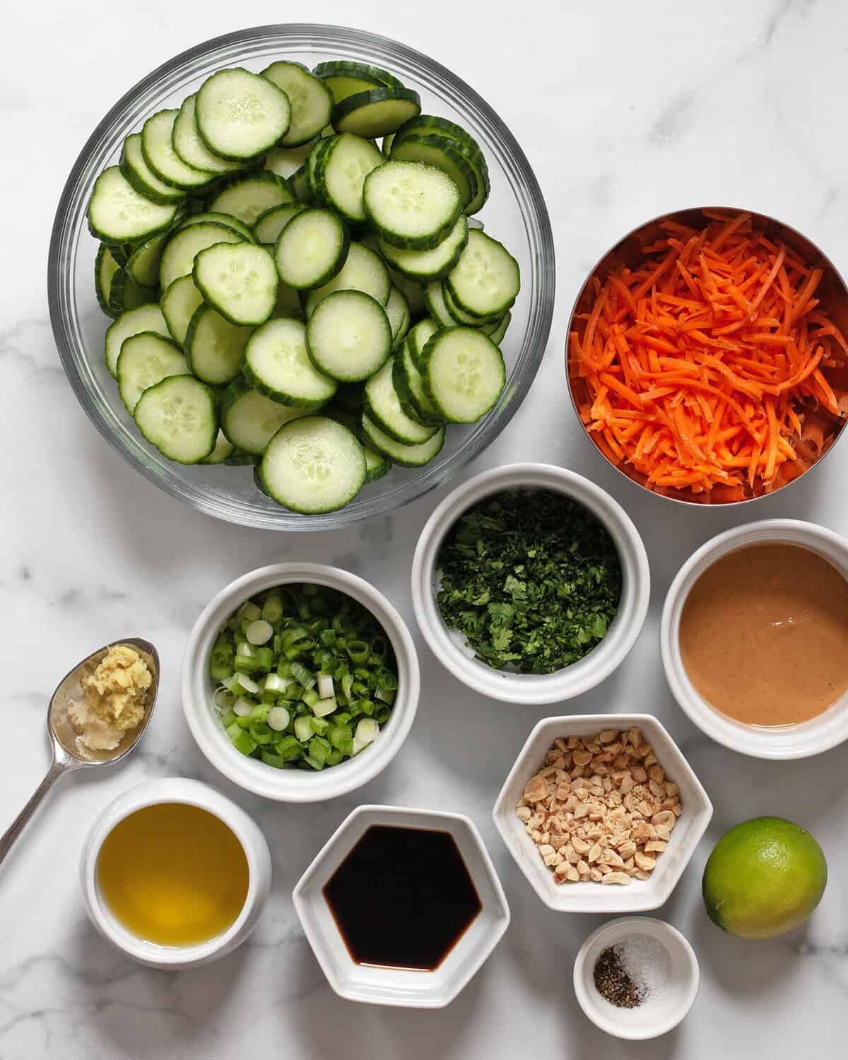 Ingredients including sliced cucumbers, shredded carrots, scallions, peanuts, peanut butter, oil, soy sauce, lime, garlic, ginger, cilantro, red pepper flakes, salt and pepper.