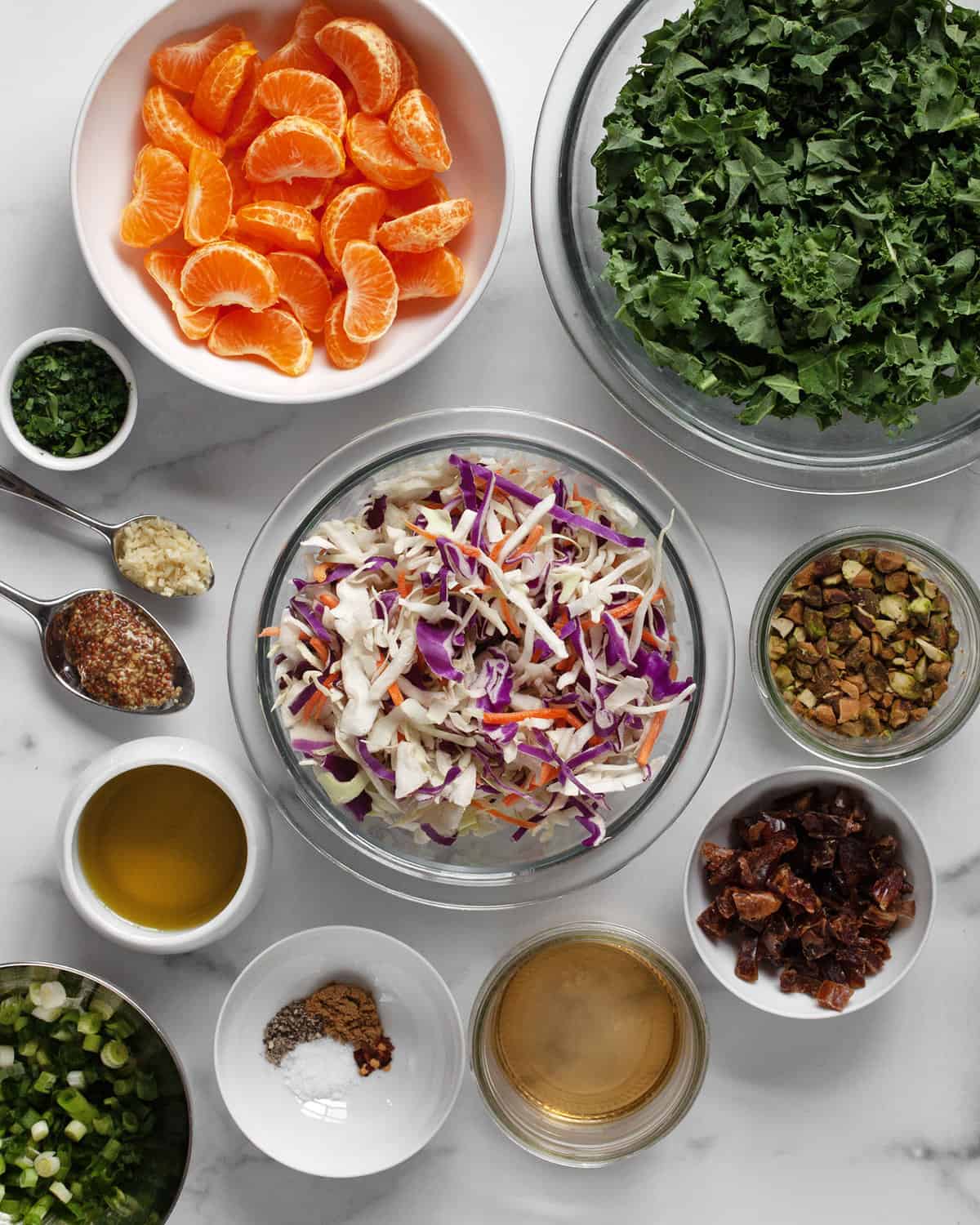 Ingredients including mandarin oranges, kale, coleslaw mix, pistachios, scallions, dates, spices, garlic, oil and vinegar.