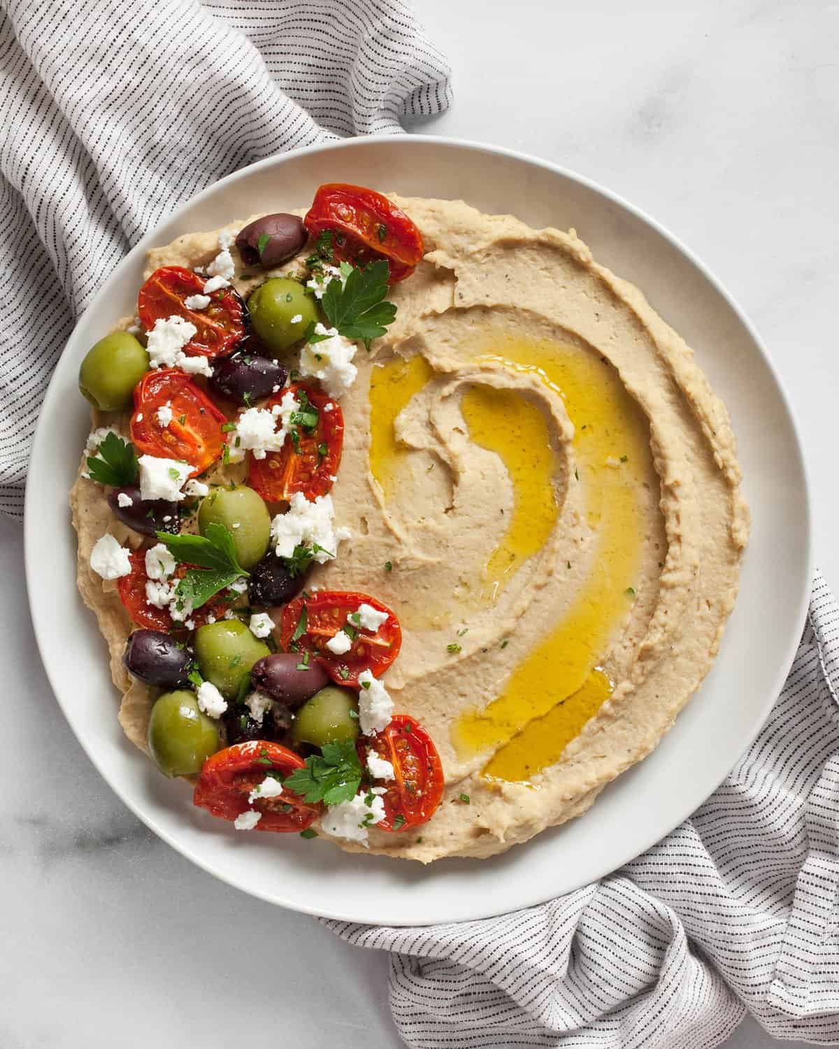 Roasted garlic hummus topped with roasted tomatoes, olives and feta spread on a plate.