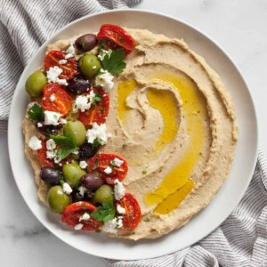 Roasted garlic hummus topped with roasted tomatoes, olives and feta spread on a plate.