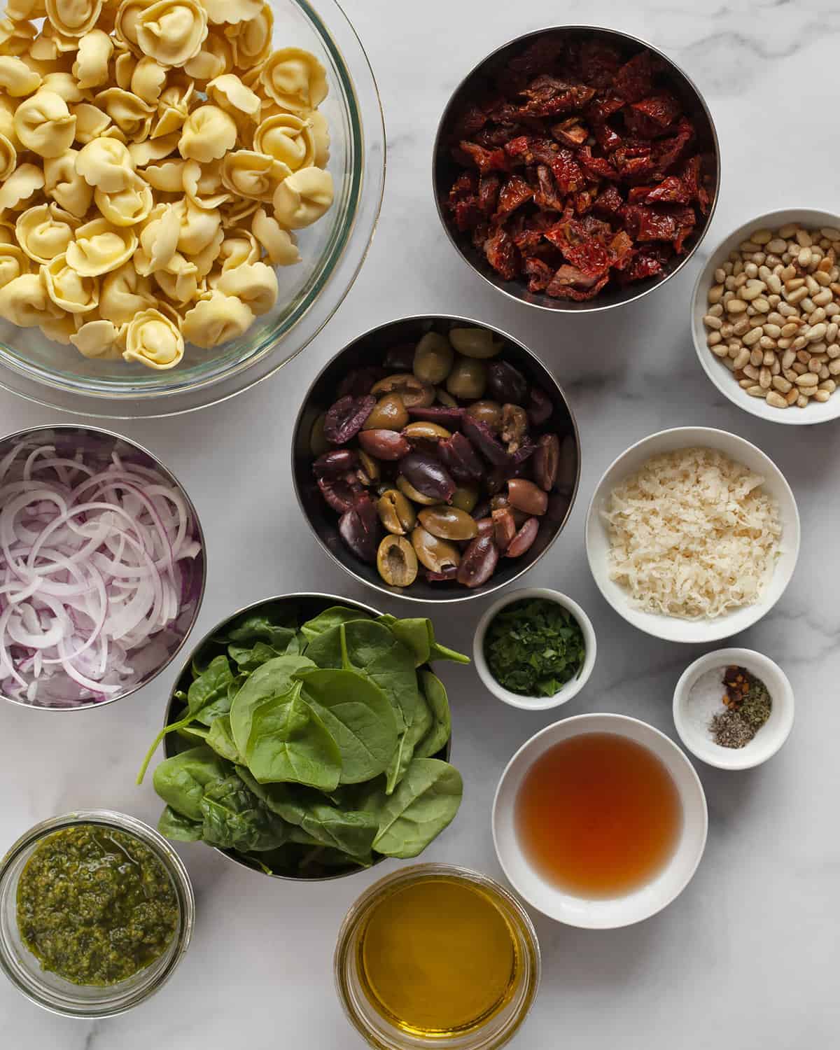 Ingredients including tortellini, sun dried tomatoes, olives, spinach, parmesan, red onions, pesto and pine nuts.