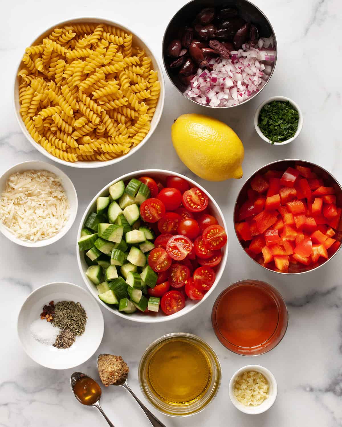 Ingredients including rotini pasta, tomatoes, peppers, cucumbers, onions, olives, vinegar, olive oil, garlic, mustard and spices.