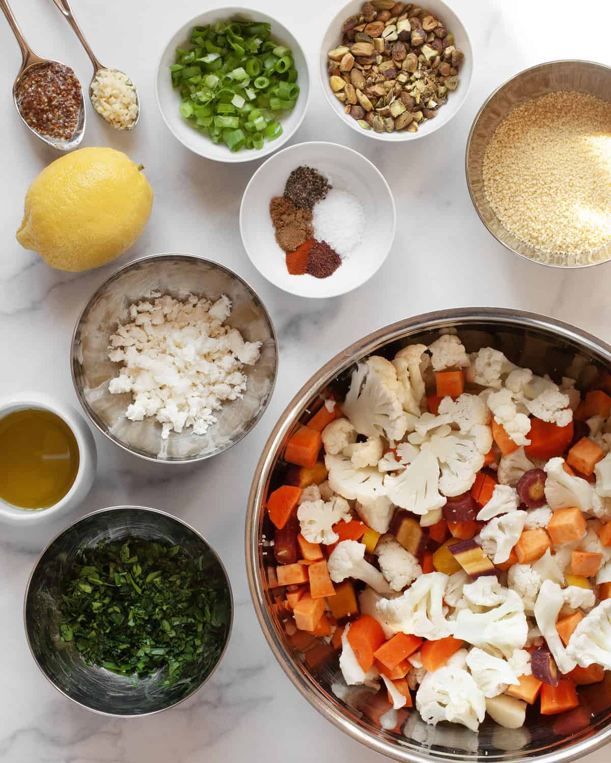 Ingredients including sweet potatoes, cauliflower, carrots, feta, scallions, parsley, pistachios, spices, couscous, lemon and oil.
