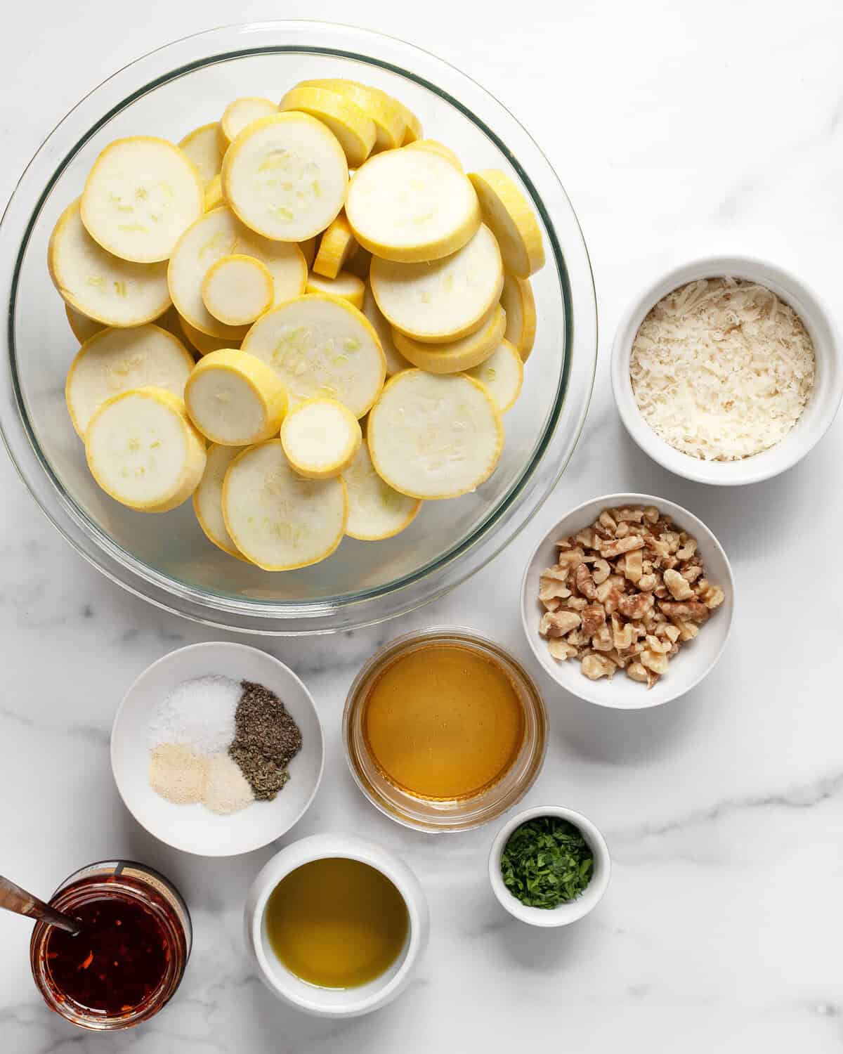 Ingredients including sliced yellow squash, parmesan, walnuts, parsley, honey, chili crisp, olive oil and dried spices.