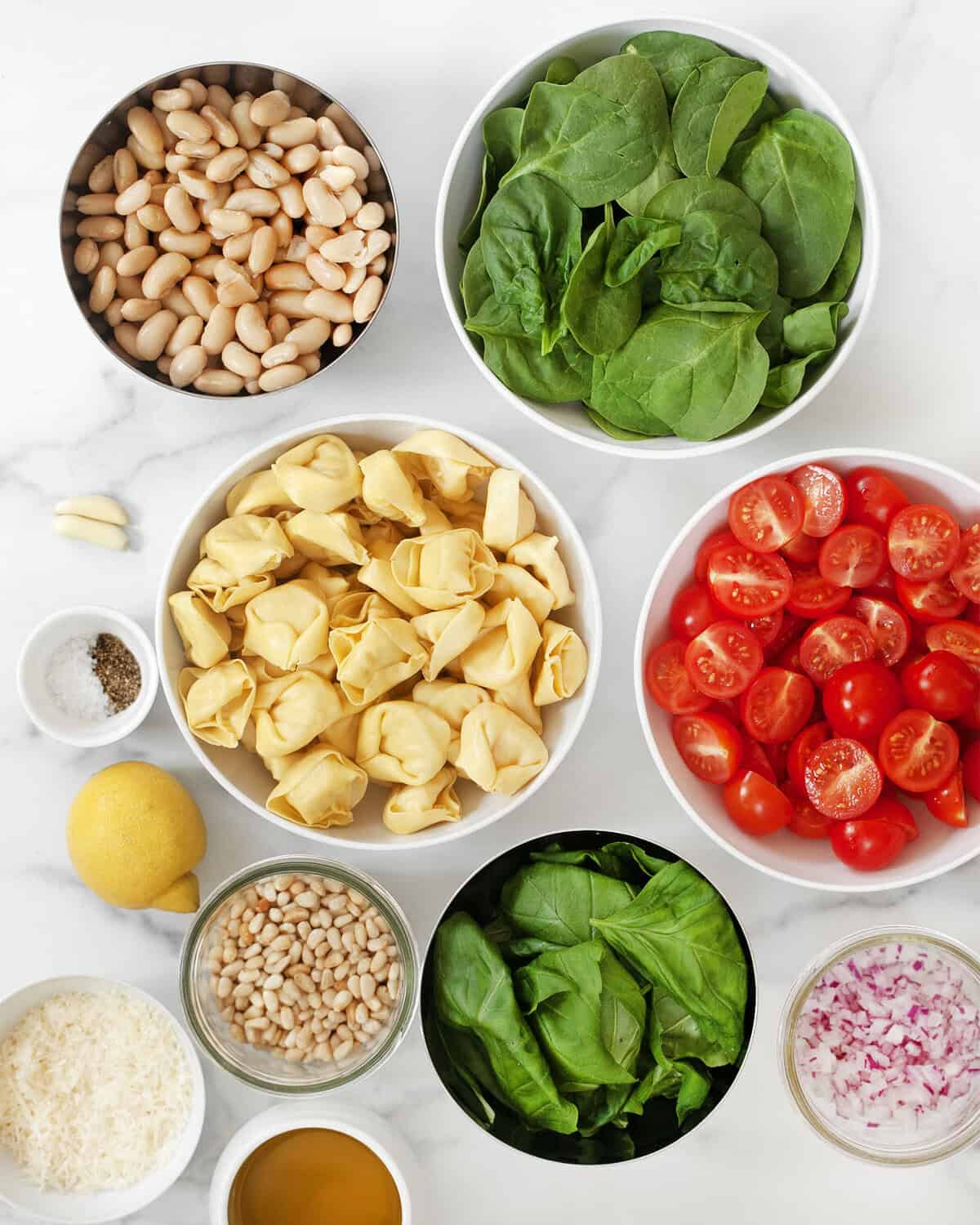 Ingredients including tortellini, white beans, tomatoes, spinach, basil, pine nuts, red onions, oil, parmesan, pine nuts, salt and pepper.