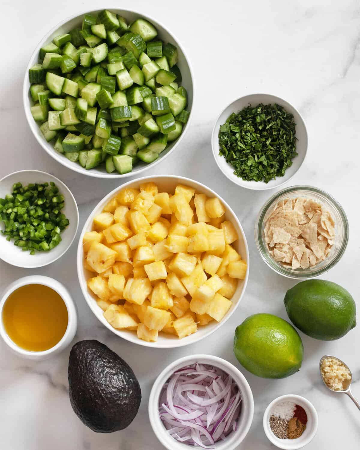 Ingredients including diced pineapple and cucumbers, lime, red onions, tortilla chips, olive oil, jalapenos, avocado, garlic and seasonings.