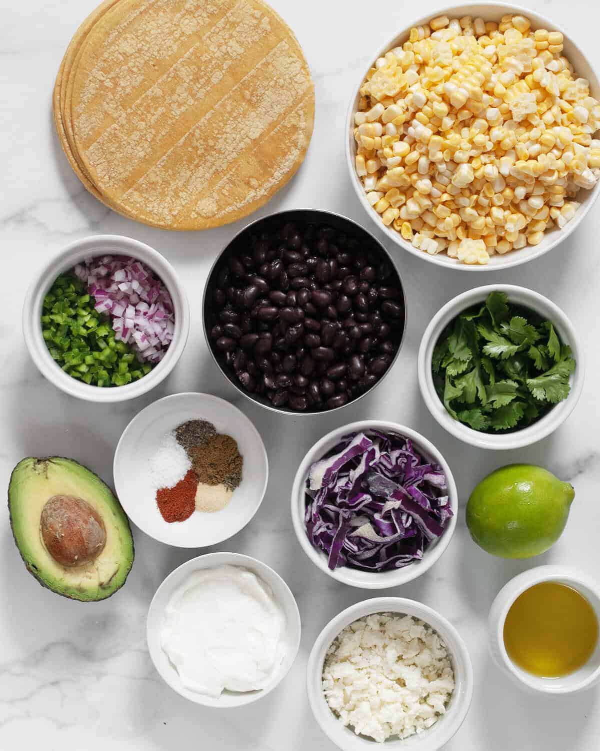 Ingredients including corn, black beans, tortillas, cilantro, red onions, jalapenos, spices, lime, feta, yogurt, avocado, olive oil and cabbage.