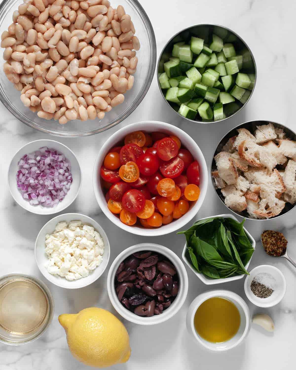 Ingredients including cannellini beans, tomatoes, cucumbers, olives, feta, red onions, basil, lemon, white wine vinegar, olive oil, salt and pepper.