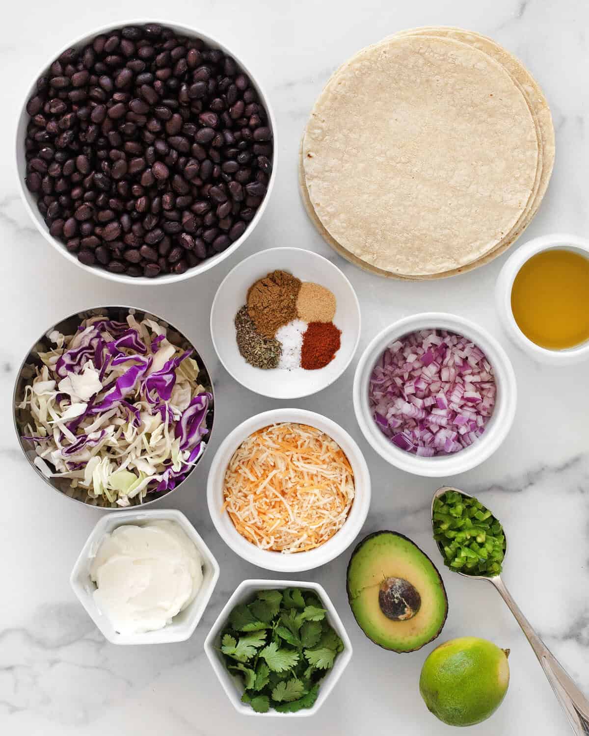 Ingredients including corn tortillas, black beans, red onions, cabbage, spices, shredded cheese, avocado, jalapenos, greek yogurt, cilantro, lime and olive oil.