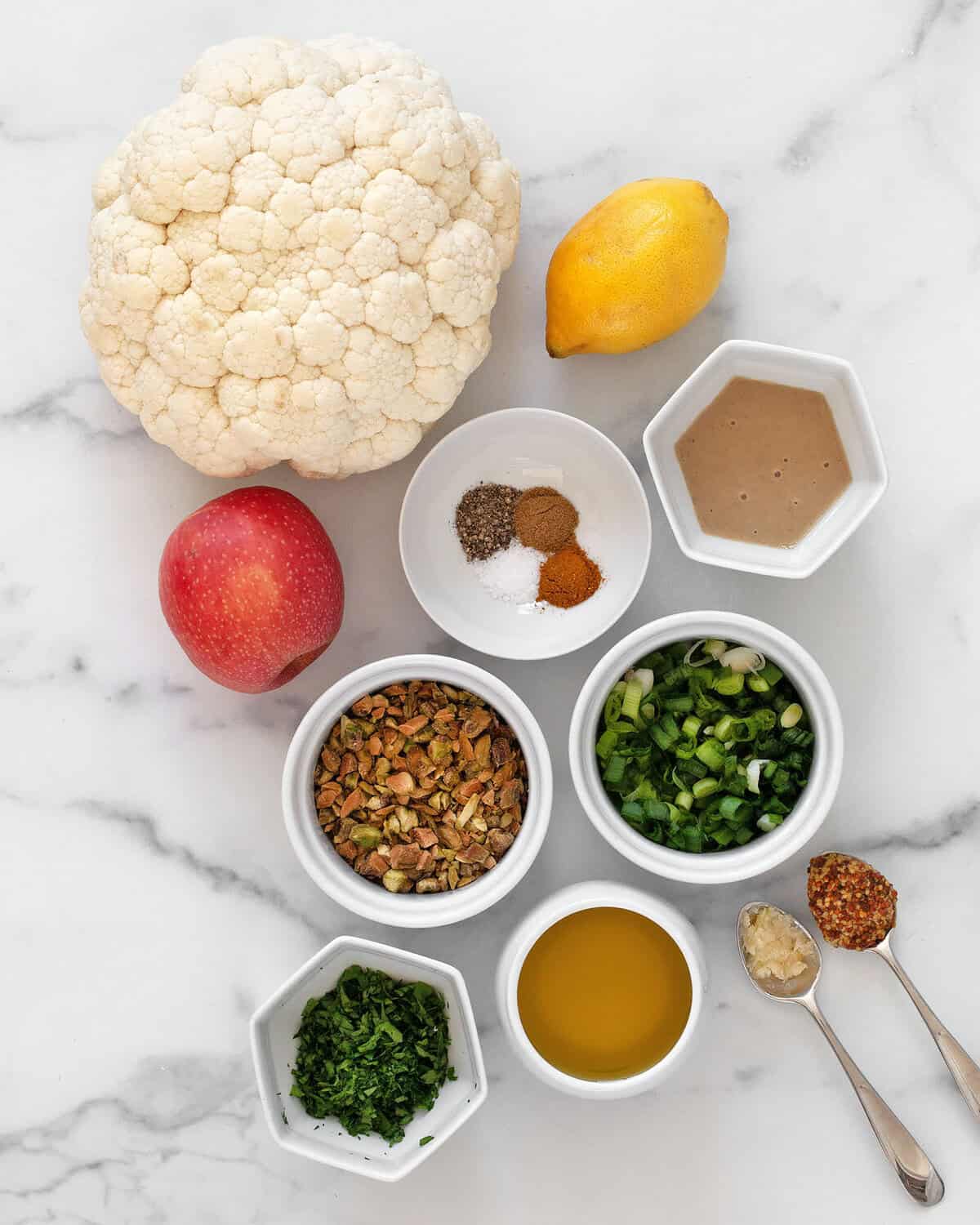 Ingredients including head of cauliflower, apple, lemon, spices, scallions, tahini, parsley, garlic, mustard and oil.