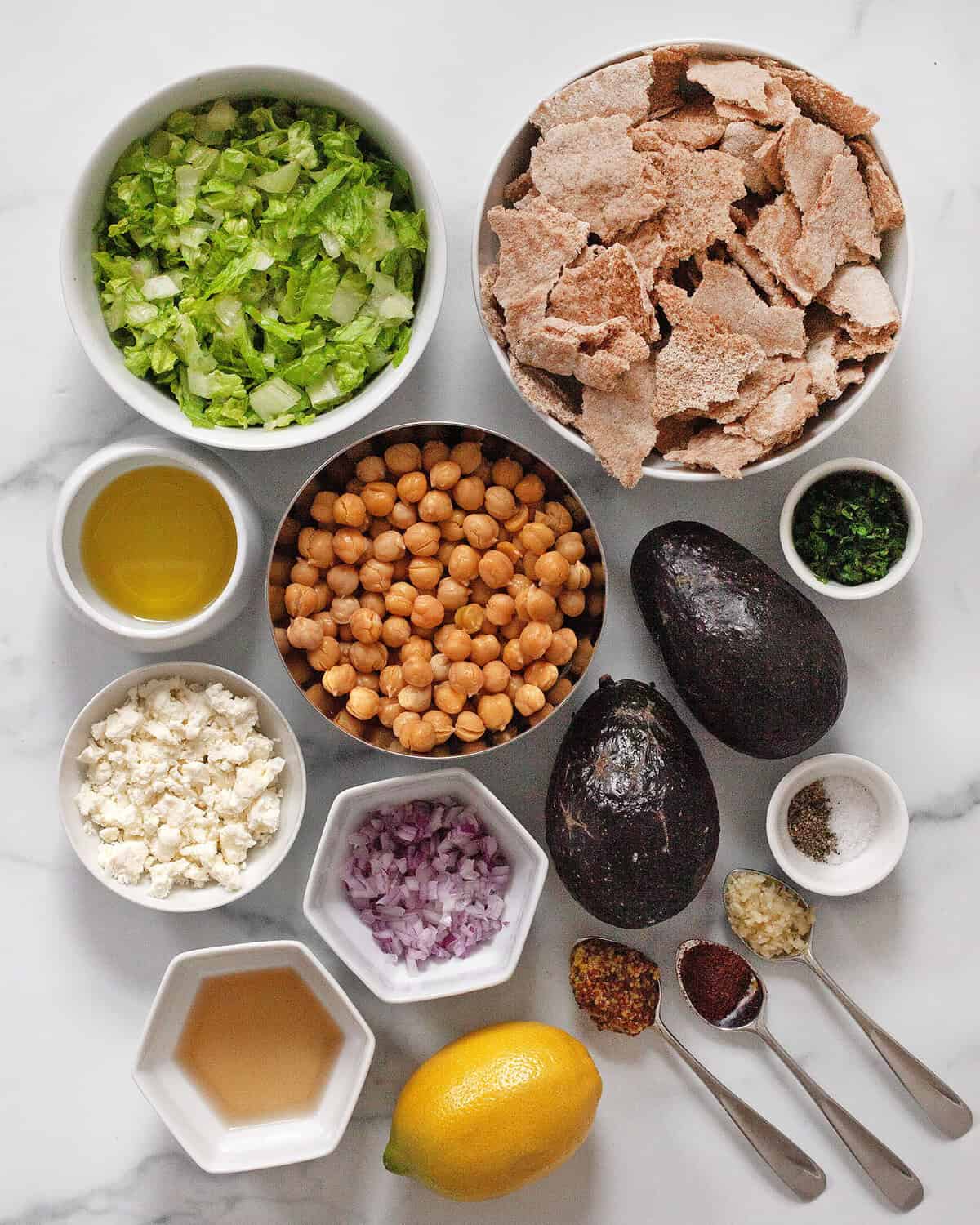 Ingredients including chickpeas, avocados, pita, feta, shredded romaine, lemon, oil, red onions, garlic, mustard, sumac, salt and pepper.
