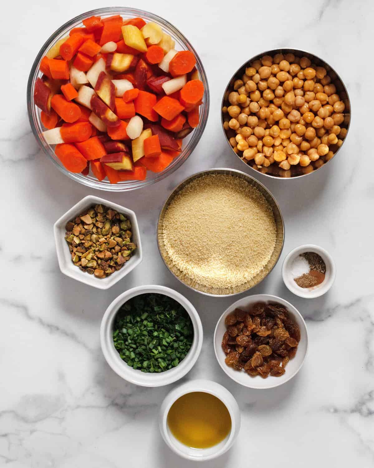 Ingredients including carrots, chickpeas, couscous, scallions, raisins. parsley & chives, pistachios, dried seasonings and olive oil.
