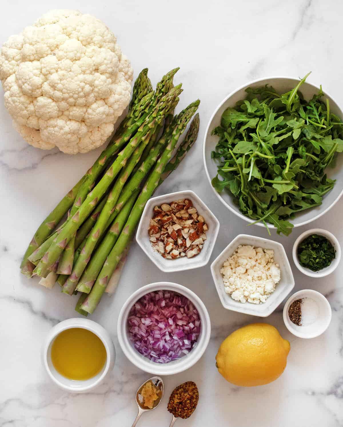 Ingredients including cauliflower, asparagus, arugula, almonds, goat cheese, red onions, lemon, olive oil, parsley, garlic, mustard, salt and pepper.