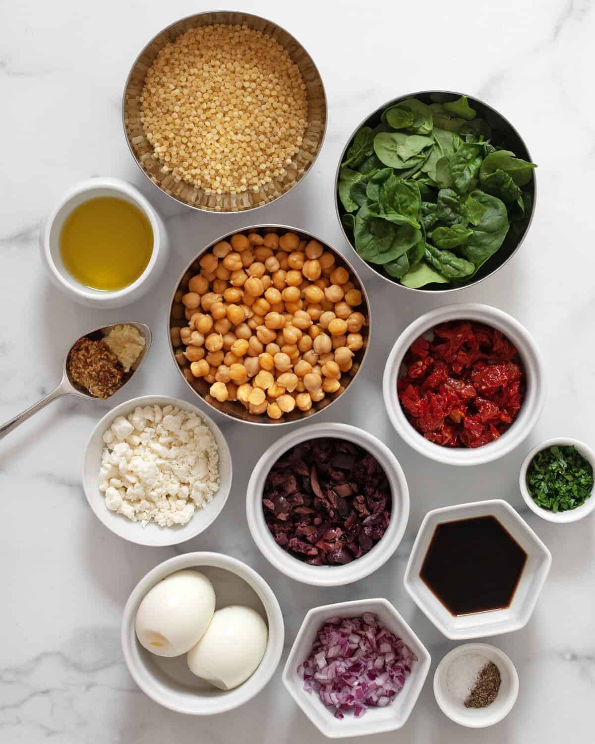 Ingredients including pearl couscous, baby spinach, chickpeas, olives, sun dried tomatoes, hard-boiled eggs, feta, olive oil, garlic, mustard, balsamic vinegar, salt and pepper.