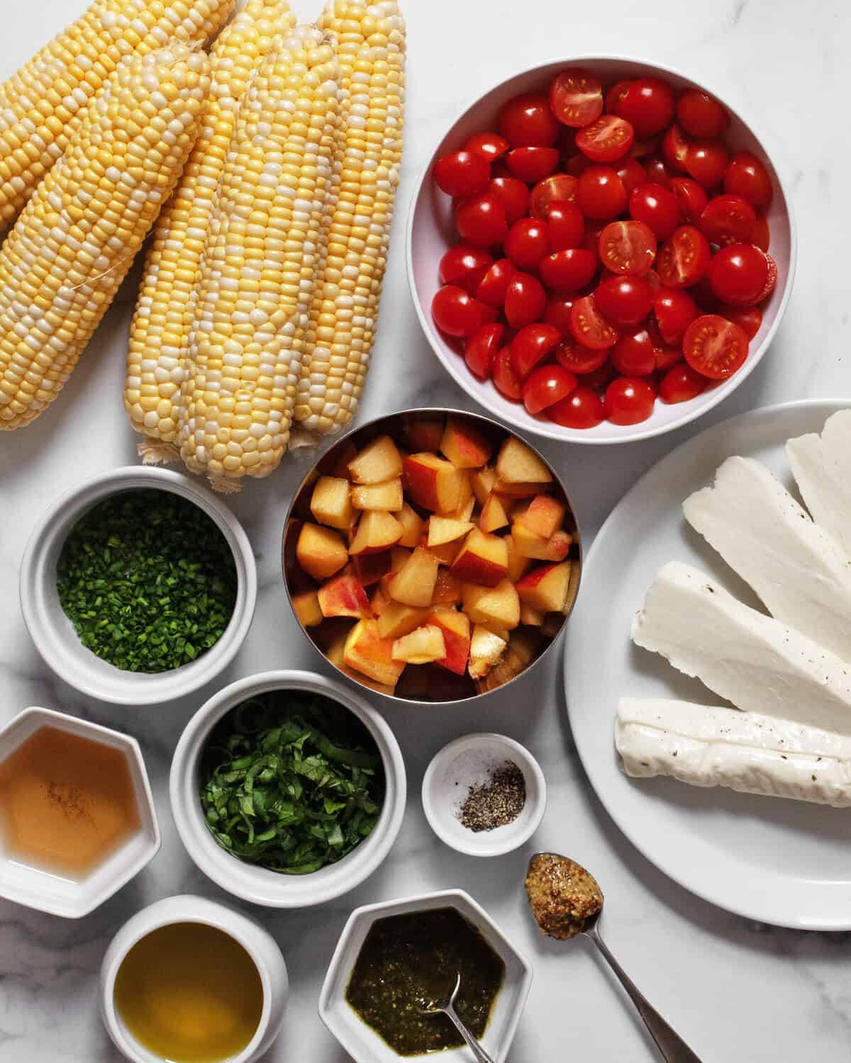 Ingredients including corn, peaches, tomatoes, halloumi, chives, basil, olive oil, mustard, white wine vinegar, pesto, salt and pepper.